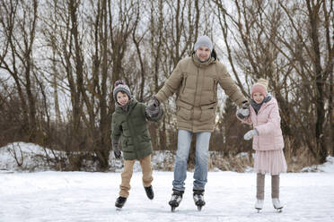 Zwei glückliche Geschwister, die zusammen mit ihrem Vater auf dem Eis Schlittschuh laufen - EYAF00858