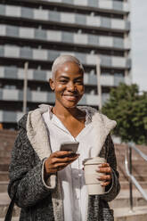 Stylish businesswoman commuting in the city, carrying smartphone and coffee cup - AFVF05077