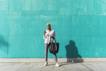 Geschäftsfrau mit Kopfhörern, die vor einer blaugrünen Wand steht und ein Smartphone benutzt - AFVF05088