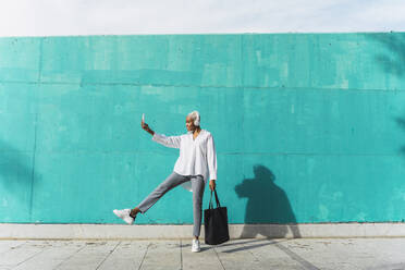 Businesswoman with headphones, standing in front of teal wall, using smartphone - AFVF05092