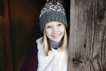 Porträt eines glücklichen Mädchens mit Wollmütze an einer Holzwand - EYAF00885