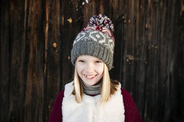 Porträt eines glücklichen Mädchens mit Wollmütze an einer Holzwand - EYAF00891