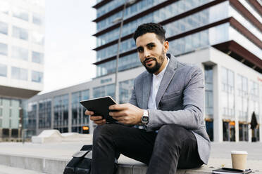 Porrait of a casual young businessman sitting on stairs in the city using tablet - JRFF03974