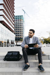 Casual young businessman sitting on stairs in the city using tablet - JRFF03975