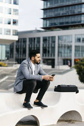 Casual young businessman sitting on a bench in the city using cell phone - JRFF03991