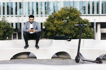 Casual young businessman sitting on a bench in the city using cell phone - JRFF03992