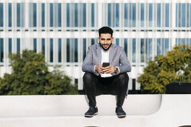 Porträt eines lässigen jungen Geschäftsmannes, der auf einer Bank in der Stadt sitzt und ein Mobiltelefon hält - JRFF03993