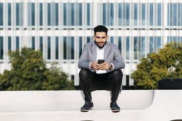 Portrait of casual young businessman sitting on a bench in the city holding cell phone - JRFF03993
