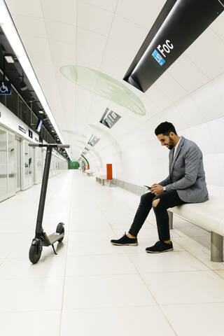 Junger Geschäftsmann mit Elektroroller und Mobiltelefon wartet in der U-Bahn-Station, lizenzfreies Stockfoto