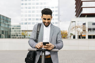 Casual young businessman with electric scooter using cell phone in the city - JRFF04020