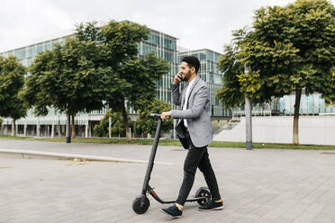 Casual young businessman with electric scooter talking on the phone in the city - JRFF04024