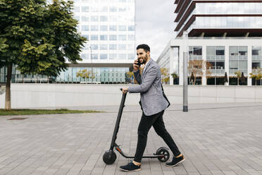 Casual young businessman with electric scooter talking on the phone in the city - JRFF04025