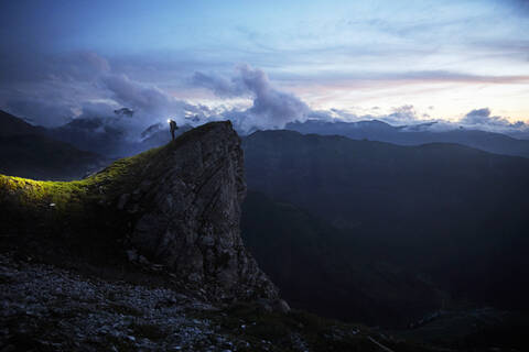 Wanderer, der in der Dämmerung auf einem Felsvorsprung steht und auf die Berge blickt, lizenzfreies Stockfoto