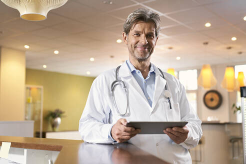 Porträt eines selbstbewussten Arztes mit Tablet am Empfang in seiner Arztpraxis - PHDF00046