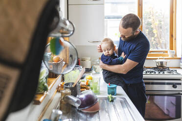 Man in the kitchen preparing food and cuddling his son - WPEF02461