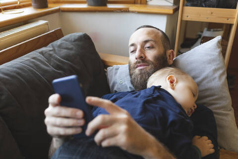 Mann schaut auf sein Handy, während sein kleiner Sohn auf dem Sofa schläft, lizenzfreies Stockfoto