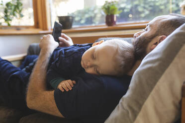 Mann schaut auf sein Handy, während sein kleiner Sohn auf dem Sofa schläft - WPEF02485