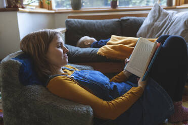 Woman reading book in living room with little son sleeping - WPEF02495