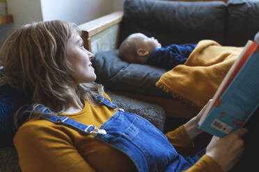 Woman reading book in living room with little son sleeping - WPEF02496