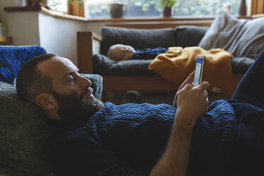 Man checking his phone while his little baby son is sleeping on the sofa - WPEF02499