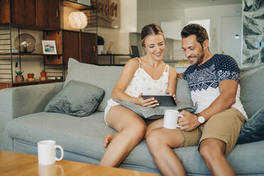 Happy relaxed couple sitting on couch in living room sharing a tablet - MPPF00458