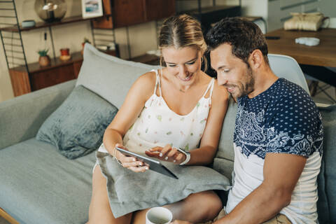 Glückliches entspanntes Paar sitzt auf der Couch im Wohnzimmer und teilt ein Tablet, lizenzfreies Stockfoto