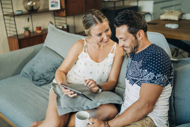 Happy relaxed couple sitting on couch in living room sharing a tablet - MPPF00460