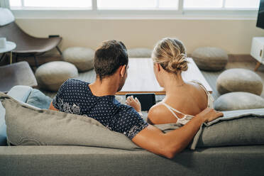 Rear view of couple sitting on couch in living room sharing a tablet - MPPF00461