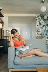 Entspannte Frau sitzt auf der Couch im Wohnzimmer und liest ein Buch - MPPF00474