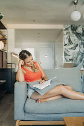 Relaxed woman sitting on couch in living room reading book - MPPF00474