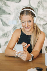 Lächelnde Frau, die mit Kopfhörern und Smartphone am Tisch sitzt - MPPF00480