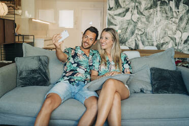 Happy couple sitting on couch in living room wearing Hawaiian shirts taking a selfie - MPPF00484