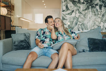 Happy couple sitting on couch in living room wearing Hawaiian shirts - MPPF00485