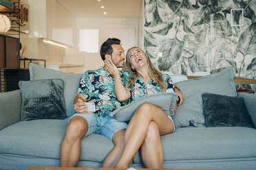 Happy couple sitting on couch in living room wearing Hawaiian shirts - MPPF00486