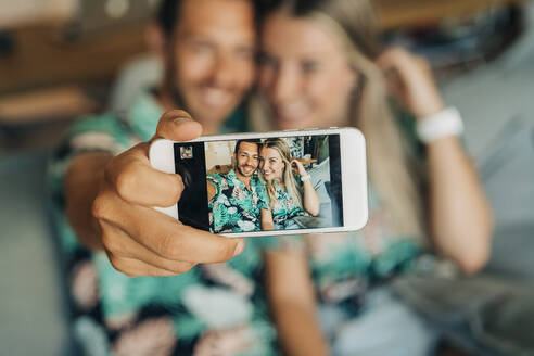 Glückliches Paar, das auf der Couch im Wohnzimmer sitzt und Hawaiihemden trägt und ein Selfie macht - MPPF00488