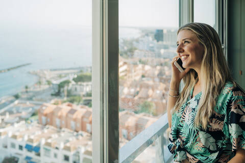 Lächelnde Frau am Telefon am Fenster in der Küstenstadt, lizenzfreies Stockfoto