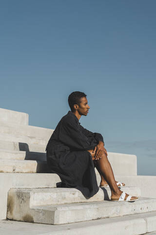 Junger Mann in schwarzem Kaftan sitzt auf einer Betontreppe unter blauem Himmel, lizenzfreies Stockfoto