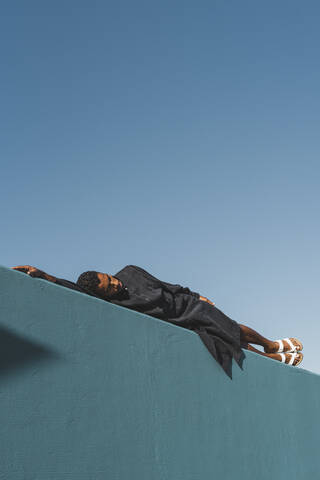 Young man wearing black kaftan lying on blue wall stock photo