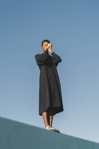 Young man wearing black kaftan standing on blue wall holding oranges in front of his face stock photo