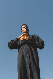 Junger Mann trägt schwarzen Kaftan unter blauem Himmel und hält Orangen - AFVF05197