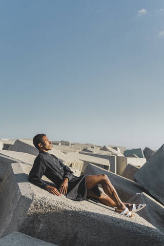Young man wearing black kaftan lying on concrete blocks under blue sky stock photo