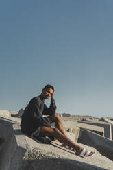 Junger Mann in schwarzem Kaftan sitzt auf Betonblöcken unter blauem Himmel - AFVF05211