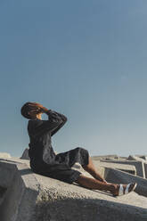 Junger Mann in schwarzem Kaftan sitzt auf Betonblöcken unter blauem Himmel und bedeckt sein Gesicht - AFVF05213