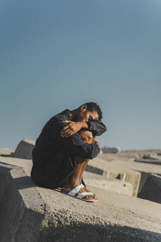 Young man wearing black kaftan sitting on concrete blocks under blue sky stock photo