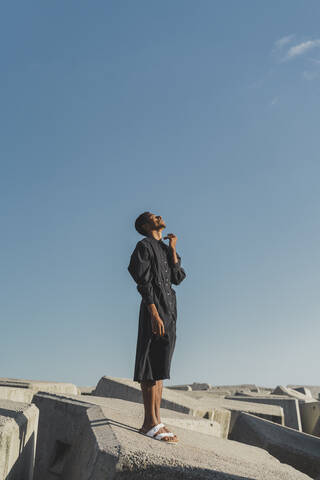 Young man wearing black kaftan standing on concrete blocks under blue sky stock photo