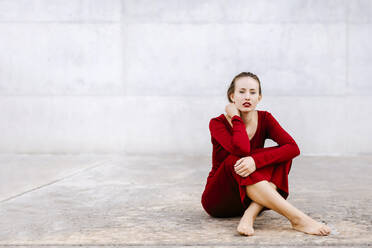 Porträt einer barfüßigen jungen Frau im roten Kleid, die im Freien auf dem Boden sitzt - TCEF00074