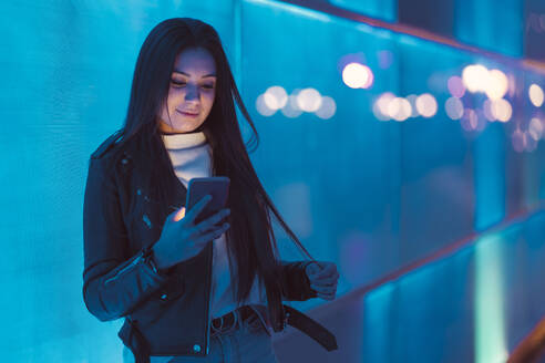 Portrait of smiling teenage girl leaning against blue glass pane looking at smartphone - DLTSF00421