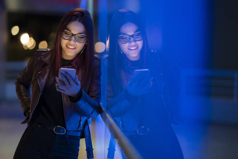 Portrait of smiling teenage girl wearing leather jacket and glasses using smartphone at night - DLTSF00423