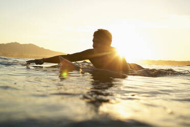 Glücklicher männlicher Surfer beim Paddeln auf dem sonnigen Meer bei Sonnenuntergang - FSIF04568