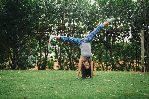Portrait fit, carefree female personal trainer doing handstand in park - FSIF04575